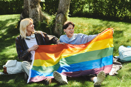 Two friends, dressed casually, sit on the grass in a park, holding a rainbow flag and laughing.の写真素材
