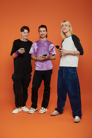 Three young queer friends stand in front of an orange background, each wearing colorful and stylish...