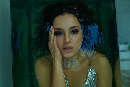 A young woman with wet hair, submerged in a blue-green pool, gazes intently through the water, her...