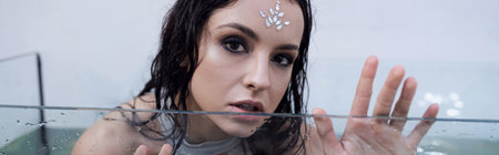 A woman with dark hair and a jeweled forehead, looks out from an aquarium, with water rippling...