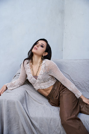 A young woman with long, dark hair wears a white knit top and brown pants while posing on a gray...