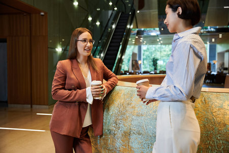 Two colleagues chat and hold coffee cups while standing by a curved reception desk in a modern office lobbyの写真素材
