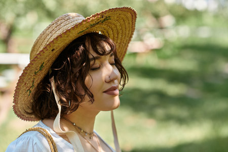A young woman with brunette hair in a straw hat and white blouse relaxes in a sunny garden, eyes closed in peaceful contemplation.の写真素材