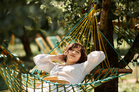 A young woman in a white dress and straw hat relaxes in a hammock, enjoying the summer sun in a lush...