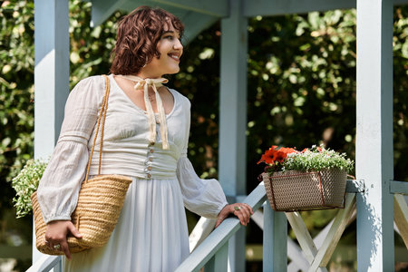 A young woman in a white dress and straw hat smiles while standing on a porch in a garden.の写真素材