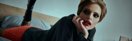 A woman in a black sweater and red pants lies on a bed, posing with a sultry look.