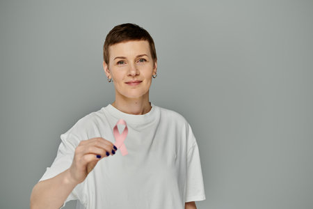 A woman with short hair, wearing casual attire, holds a pink ribbon in front of a grey background.の写真素材