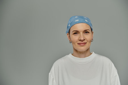 A woman with short hair wears a blue bandana and smiles softly, standing against a grey backdrop.