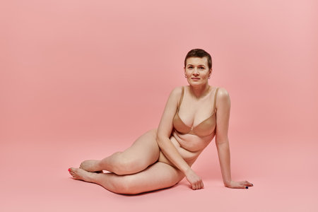 A woman with short hair wearing a bra sits on a pink background, posing for breast cancer awareness.の写真素材