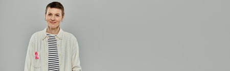 Woman with short hair and pink ribbon pinned to shirt, symbolizing breast cancer awareness, looks hopeful at cameraの写真素材