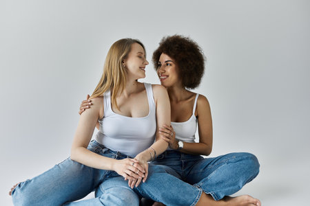 An interracial lesbian couple in white tank tops and jeans sits together on a grey background, smiling and looking at each other.の写真素材