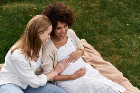 A pregnant Black woman sits on a blanket in the grass, her partner lovingly cradles her belly.の写真素材