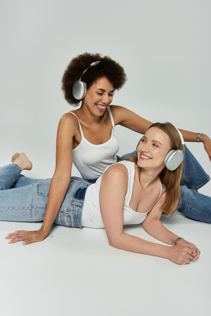 A Black and White lesbian couple in tank tops and jeans pose on a grey background, listening to music on headphonesの写真素材