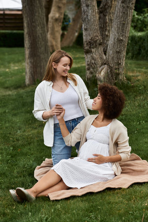 A pregnant woman sits on a blanket in a grassy park, her partner lovingly touches her hand while...