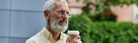 A man with a white beard, wearing glasses, holds a cup of coffee outdoors near a modern building.