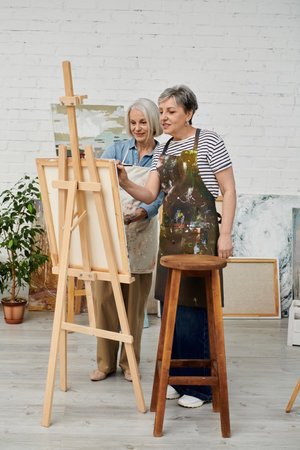 Two women, wearing paint-splattered aprons, work together on a canvas in a bright art studio.の写真素材
