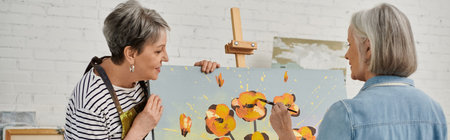 Two women paint in a studio, one focused on brushstrokes while the other watches, both in casual clothingの写真素材