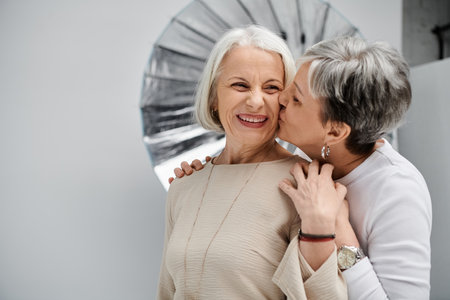 A mature lesbian couple poses for a portrait in a photo studio, filled with affection as one kisses the other on the cheek.の写真素材