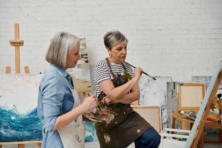 Two mature women, wearing painting smocks, stand in an art studio, one holding a paintbrush, the other holding a palette.の写真素材