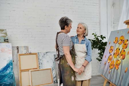 Two women in an art studio, dressed in painting aprons, hold hands and look at each other lovingly.