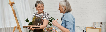 Two mature women stand in an art studio, one holding a paintbrush and palette, while the other...
