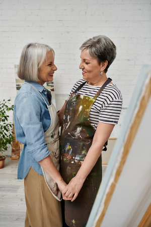 A mature lesbian couple shares a loving moment in their art studio, their hands intertwined as they paint and learn together.の写真素材