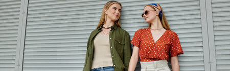 Two young women stand near wall, smiling and looking at each other.