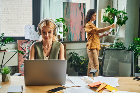 Two women in business attire work in a modern office - one on a laptop, the other reviewing documents in the backgroundの写真素材