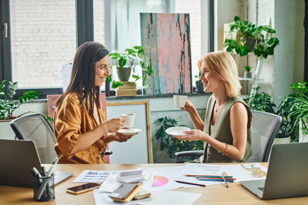 Two women, dressed in smart casual attire, enjoy a coffee break while working on a project in a contemporary office setting.の写真素材