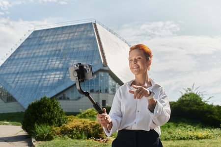 A woman records a video on her phone while standing in front of a modern campus building.の写真素材