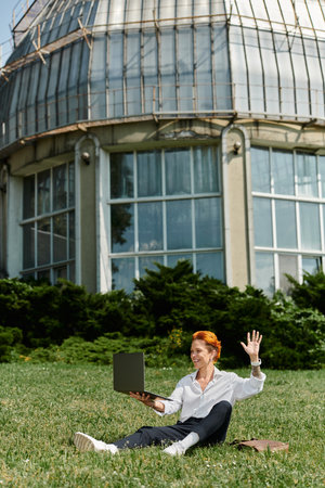 A teacher with red hair sits on a grassy lawn outside a building, waving to a laptop screen.の写真素材