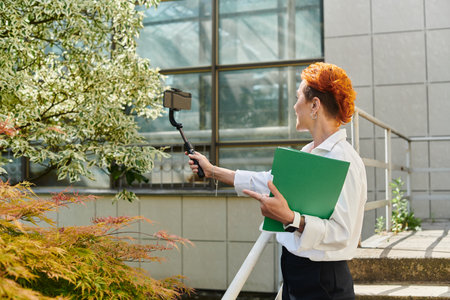 Red-haired woman takes a selfie with a phone on a selfie stick while holding a green folder