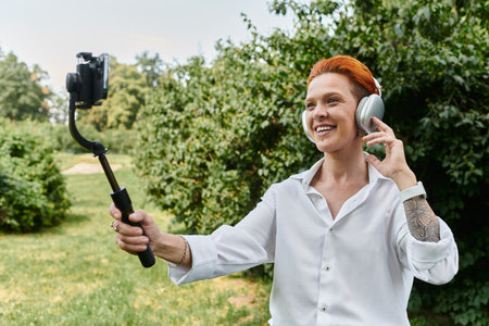 A woman in a white shirt smiles while recording a video with a selfie stick outdoors.