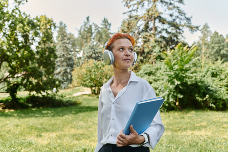 Woman in white shirt, headphones, holding blue folder, walks through green campus