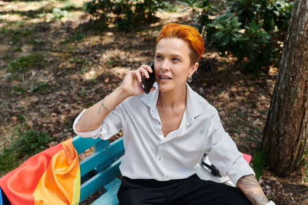 A teacher with red hair sits on a bench on a campus, talking on the phone.