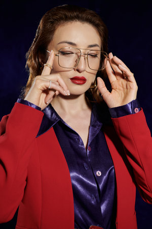 A stylish Asian woman in a red blazer and purple shirt adjusts her glasses, lost in thought.の写真素材