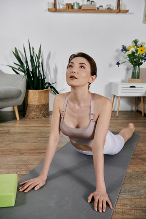 A beautiful Asian woman in a crop top performs a yoga pose on a mat in her modern apartment, her...