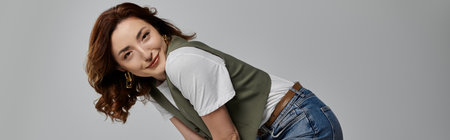 A beautiful Asian woman in a green vest, white t-shirt, and jeans poses against a grey backdrop, her brown hair framing her smiling face.の写真素材