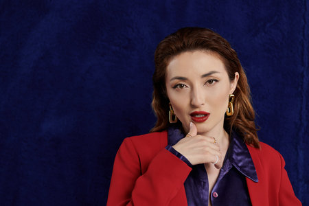 Stylish Asian woman in red suit and silk shirt poses confidently against dark blue background with bold makeup, creating striking imageの写真素材