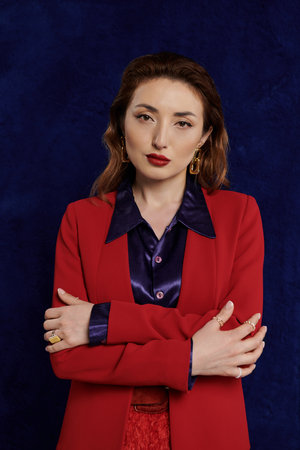 An Asian woman in a red suit and silk shirt poses with confidence against a dark background, her gaze captivating.の写真素材