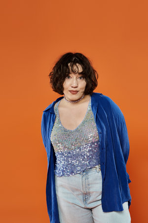 A woman in a shimmering top and a bright blue jacket stands against a vibrant orange backdrop.