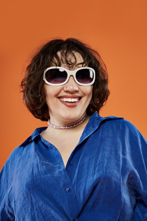 A woman in sunglasses and a blue shirt smiles broadly against an orange background.