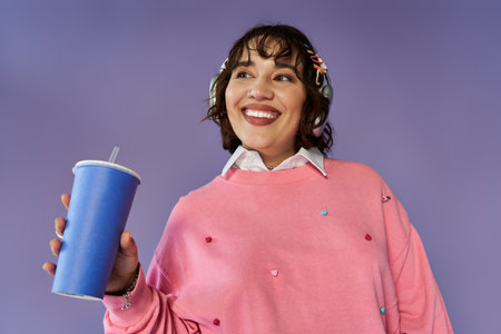 A woman in a pink sweatshirt and headphones holds a blue cup with a straw, smiling brightly.の写真素材