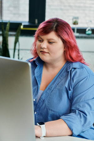 A plus size woman with pink hair works intently on a laptop.の写真素材