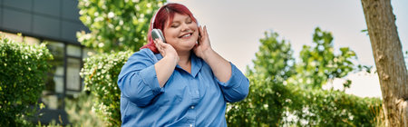 A plus size woman with red hair smiles while listening to music in a beautiful garden.