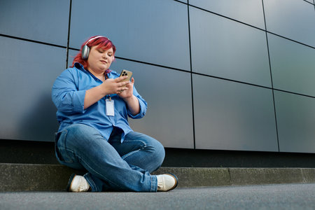 A plus size woman sits on a curb in front of a modern building, lost in her phone.