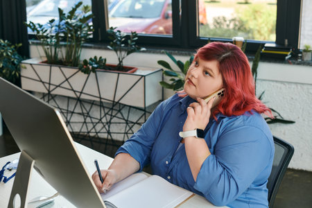 A plus size woman sits at a desk, taking notes while on a phone call.の写真素材