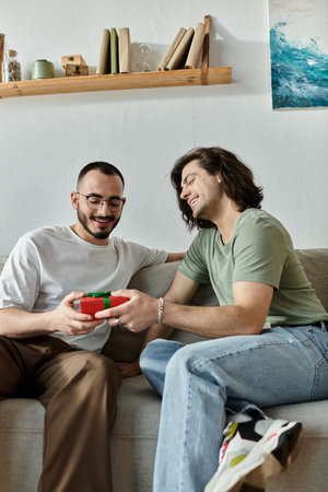 A gay couple sits on a couch, one presenting a gift to the other while both smile happily.