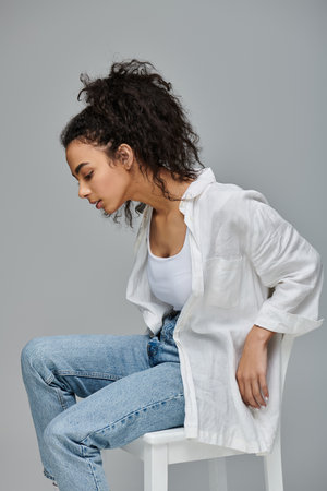 A young woman with curly hair sits on a white stool, wearing a white shirt and blue jeans, her gaze directed down thoughtfully.の写真素材