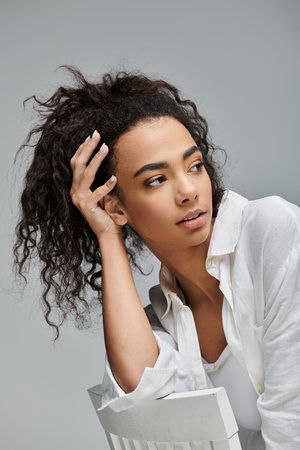 A young woman with curly hair poses thoughtfully against a gray backdrop, her gaze directed away...